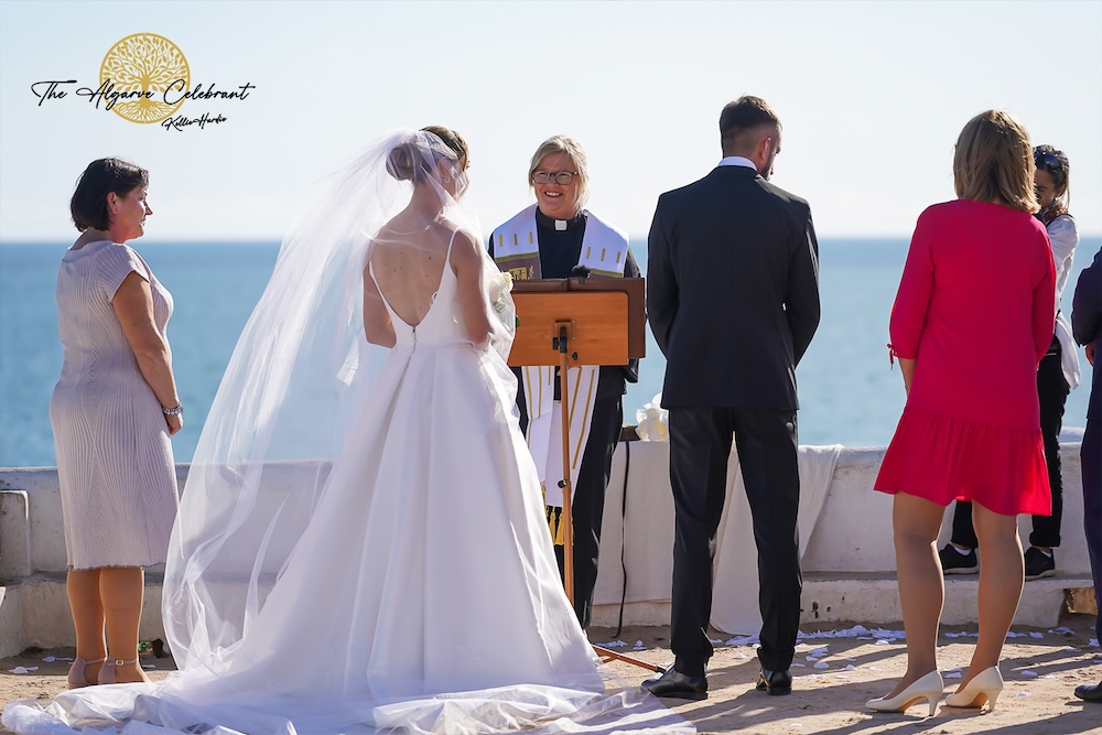 The Couple Outside the Chapel: Daniel and Magdalena standing together outside Senhora da Rocha chapel, holding hands and smiling against the backdrop of the ocean.